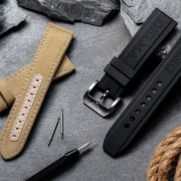 A collection of tech-savvy watch straps and accessories, featuring a tan fabric strap and a black rubber strap, displayed on a textured stone background with tools