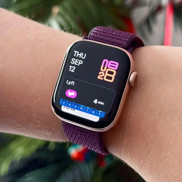 A close-up of a wrist wearing a stylish smartwatch with a purple strap, displaying the date and time, set against a backdrop of greenery and festive decorations