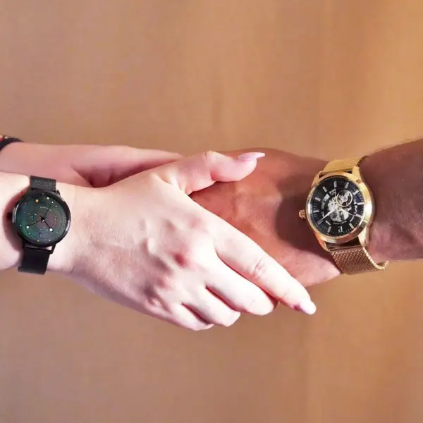 Two hands reaching out to each other, one wearing a black smartwatch and the other a gold watch, against a neutral background