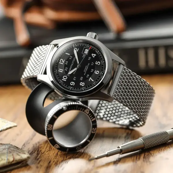 A sleek smartwatch with a metal mesh strap sits on a wooden surface, accompanied by a circular tool and a black bezel, highlighting its stylish design and functionality