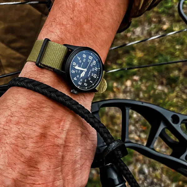 A close-up of a hand wearing a black outdoor watch with a green strap, gripping a bowstring, showcasing the watch's durability for outdoor adventures