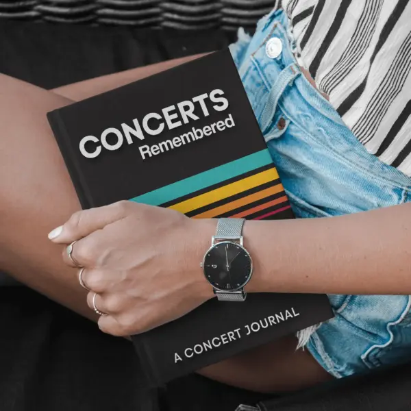 A person holds a concert journal titled "Concerts Remembered" while wearing a stylish watch and casual denim shorts, showcasing a trendy concert outfit