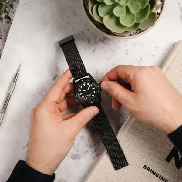 A person adjusts a quick release watch strap on a black watch, surrounded by a succulent plant, a notebook, and a pen on a marble surface