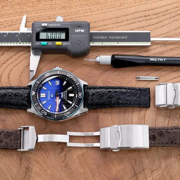 A close-up of a watch with a blue dial, surrounded by tools and two watch straps. A caliper measures the strap's width, showcasing a deployant clasp installation process