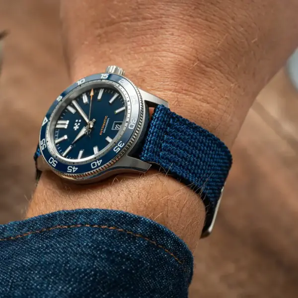 A close-up of a wrist wearing a blue nylon watch strap, showcasing a stylish watch with a blue dial. The background features a plant and a coffee cup on a wooden table