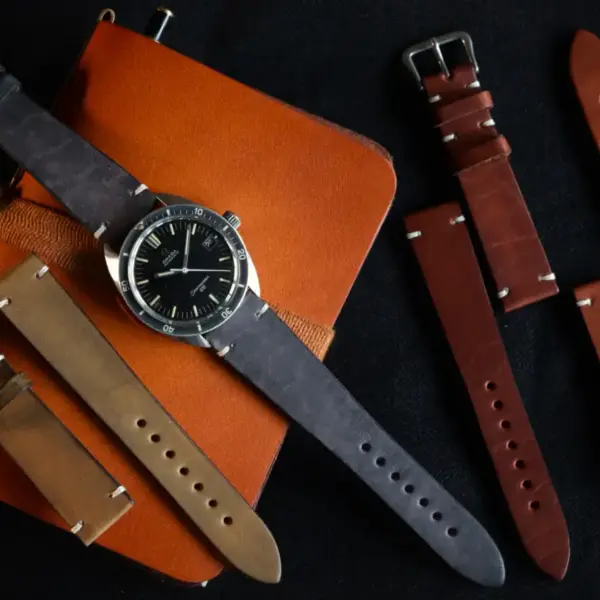 A stylish watch with a black face is placed on a brown leather notebook, surrounded by various vintage leather watch straps in shades of brown and tan