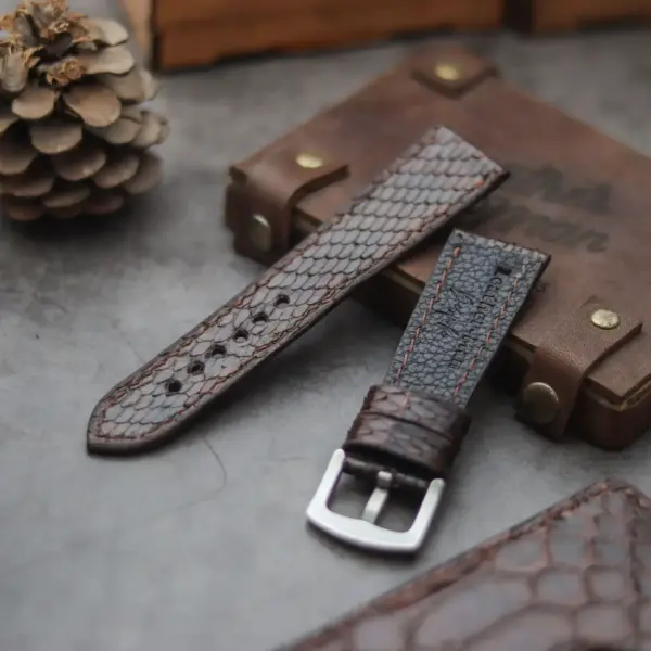 A close-up of a snake skin watch strap placed on a textured surface, alongside a wooden box and a pinecone, highlighting its unique design and craftsmanship