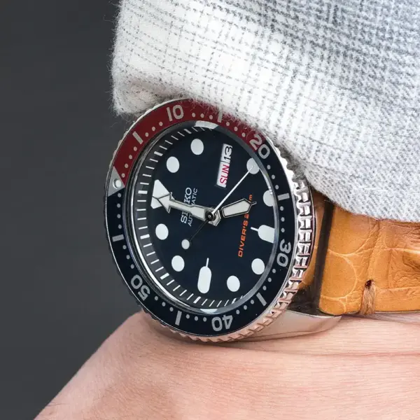 A close-up of a wrist wearing a watch with a black dial and red accents, paired with a stylish crocodile leather strap. The background is softly blurred to highlight the watch