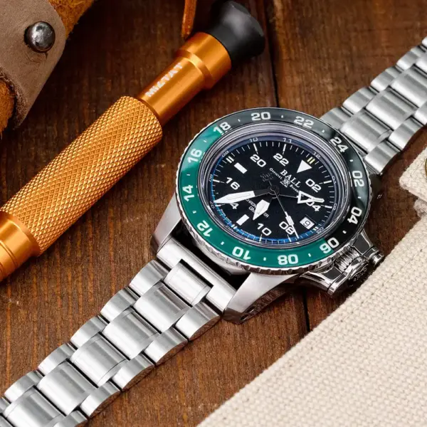 A close-up of a metal watch with a black dial and green bezel, placed on a wooden surface alongside a tool and fabric, highlighting watch styling and accessories