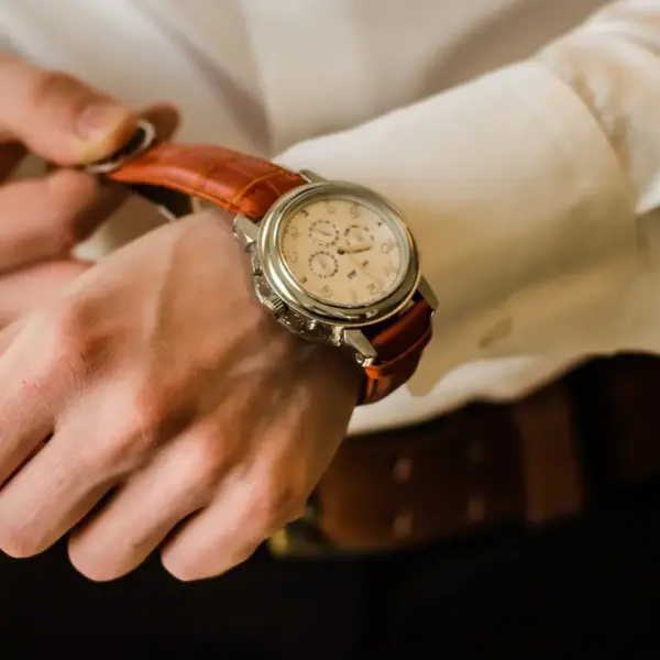 A close-up of a person adjusting a stylish watch with a brown leather strap on their wrist, dressed in a white shirt and dark pants, showcasing a fashionable accessory