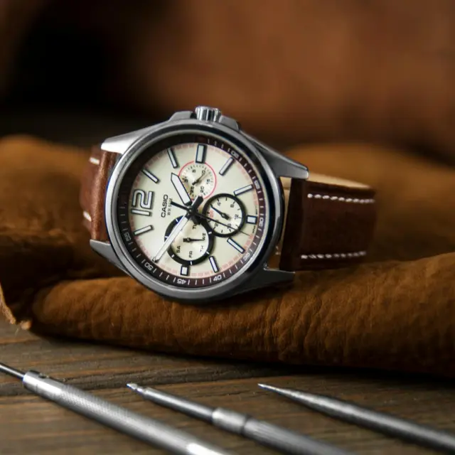 Stylish round wristwatch with chronograph and brown strap placed on leather material near repair tools in modern workshop