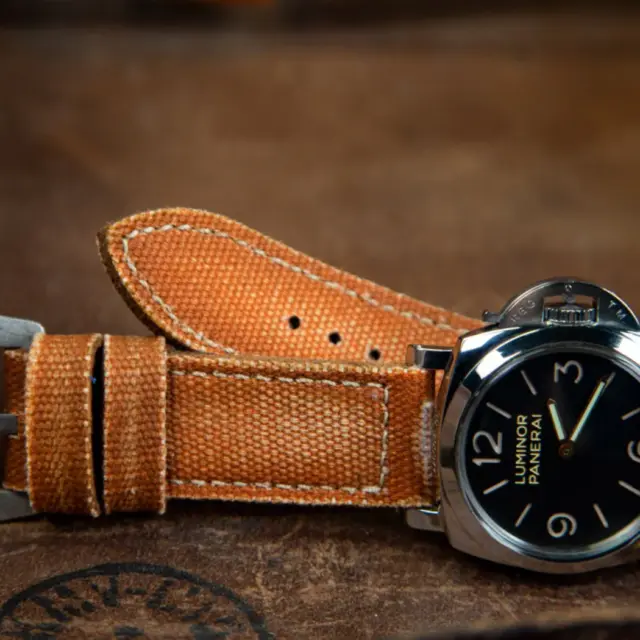 A vintage watch with a black dial and silver casing rests on a brown leather surface, showcasing a worn orange strap that adds a nostalgic touch