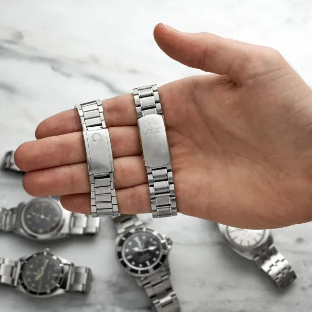 A hand holds two classic silver watch bracelets, surrounded by various watches on a marble surface, showcasing the timeless appeal of watch accessories