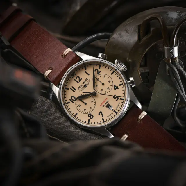 A vintage pilot watch with a cream dial and brown leather strap, resting on aviation gear, highlighting the connection between aviation and watch design